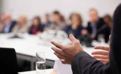 A business-man's hands gesture. In the background are people gathered around a conference 